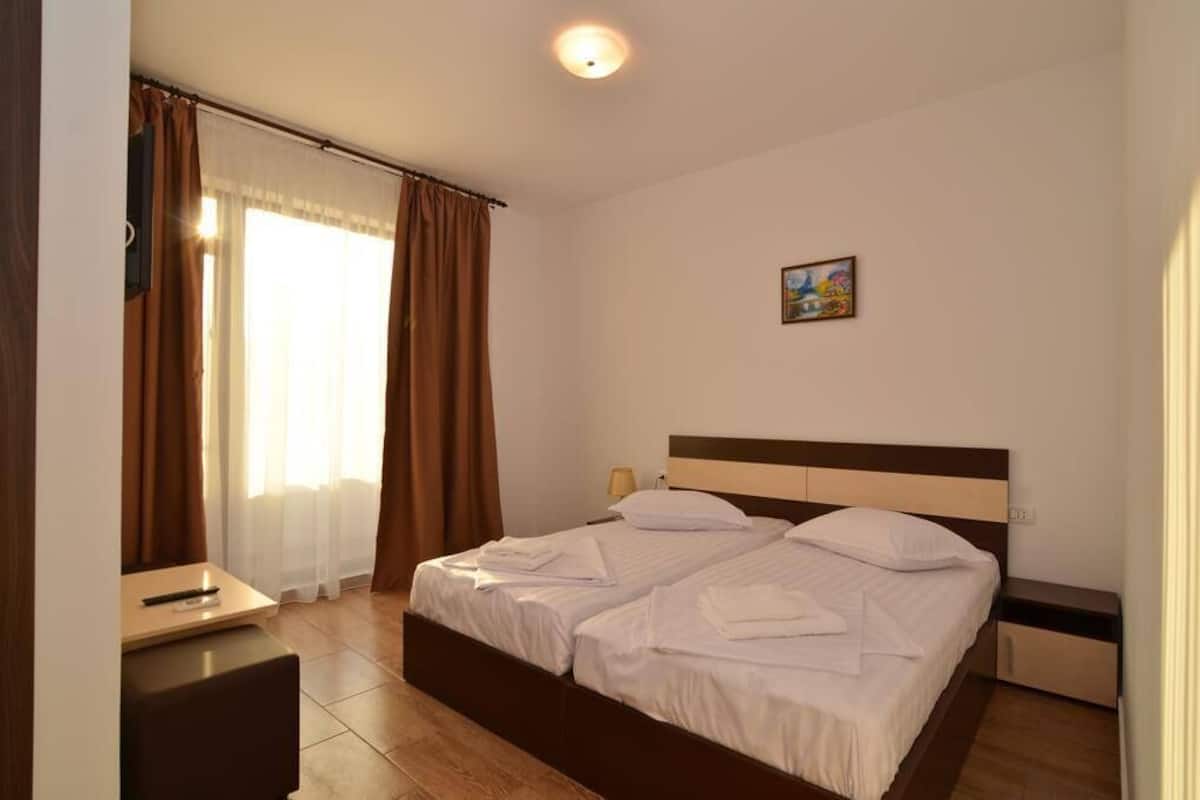 A hotel room with a double bed, a bedside table, a lamp, a window with curtains, and a painting on the wall.