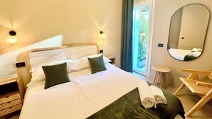 A hotel room with a bed, a round mirror, a small round table, and a view of greenery through a window.