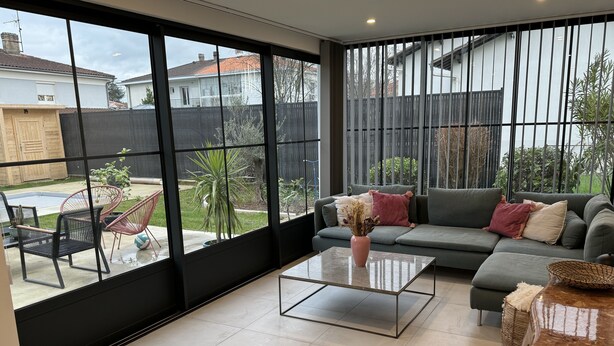 A modern living room with a grey sofa, a glass coffee table, and a view of a backyard through large windows. - Maison d Isaure (Bergerac)
