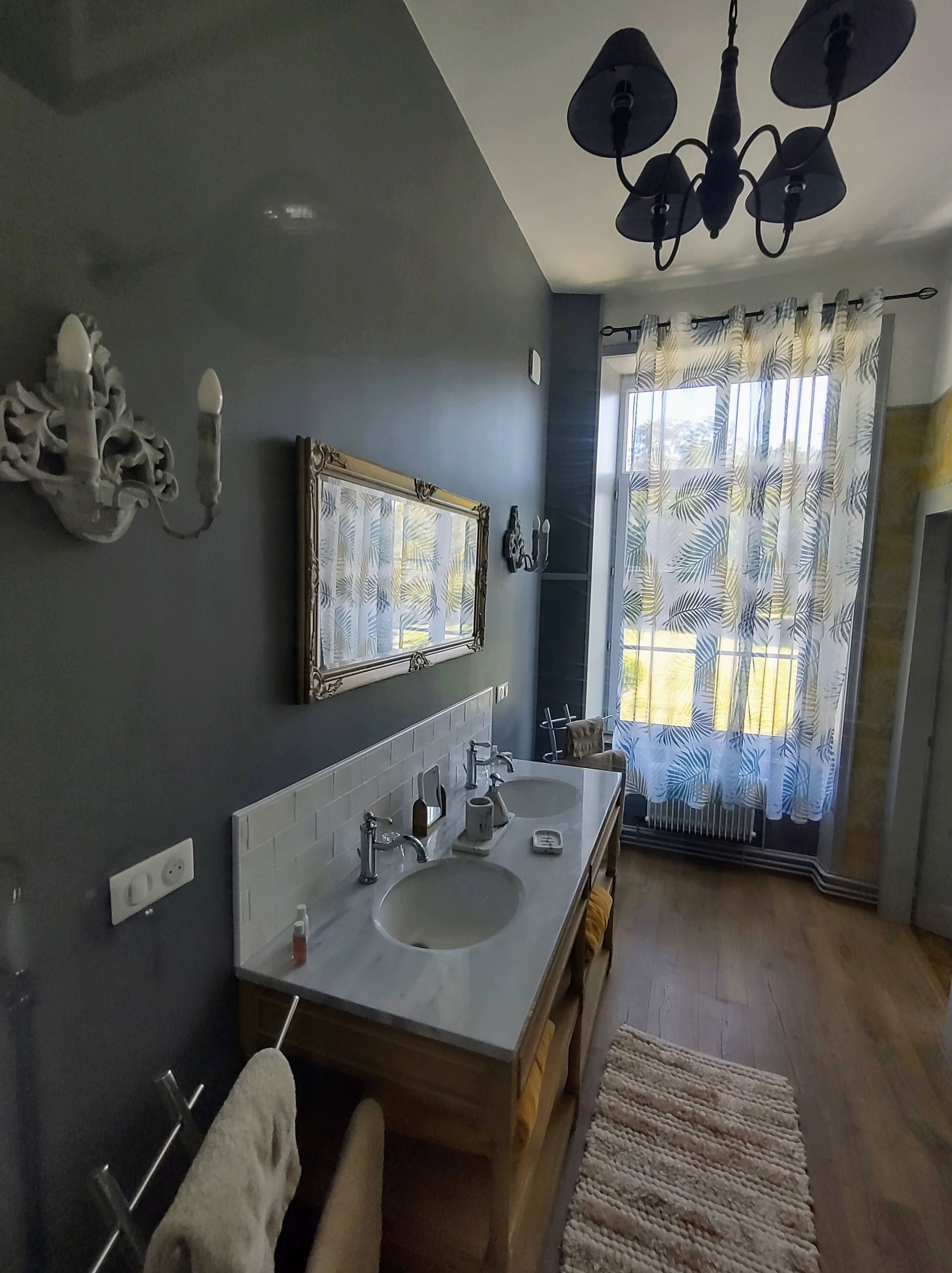 Un baño con lavamanos doble, espejo, candelabro y ventana con cortinas.