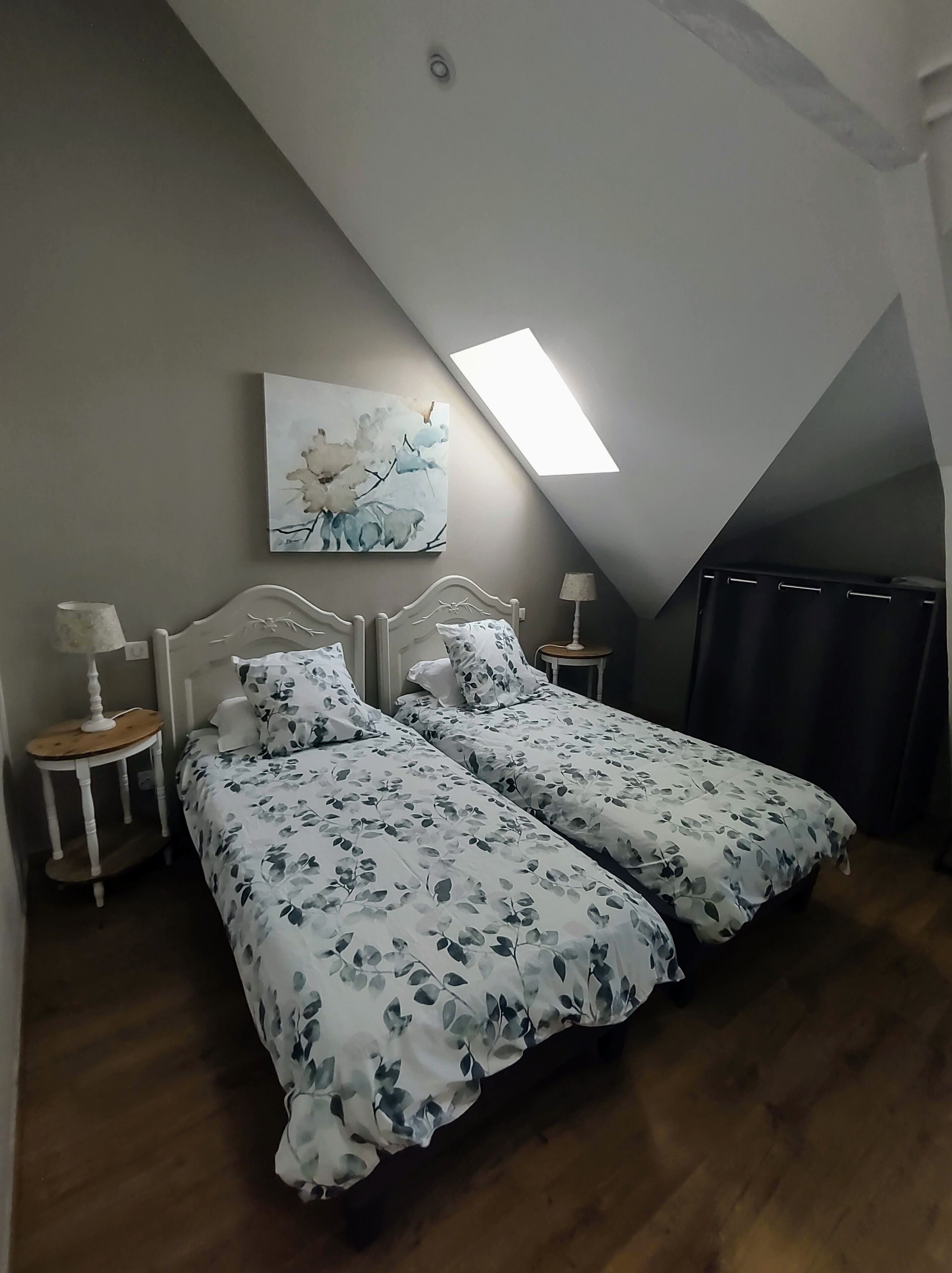 Une chambre avec deux lits, une lucarne, un tableau au mur et une petite table avec une lampe.