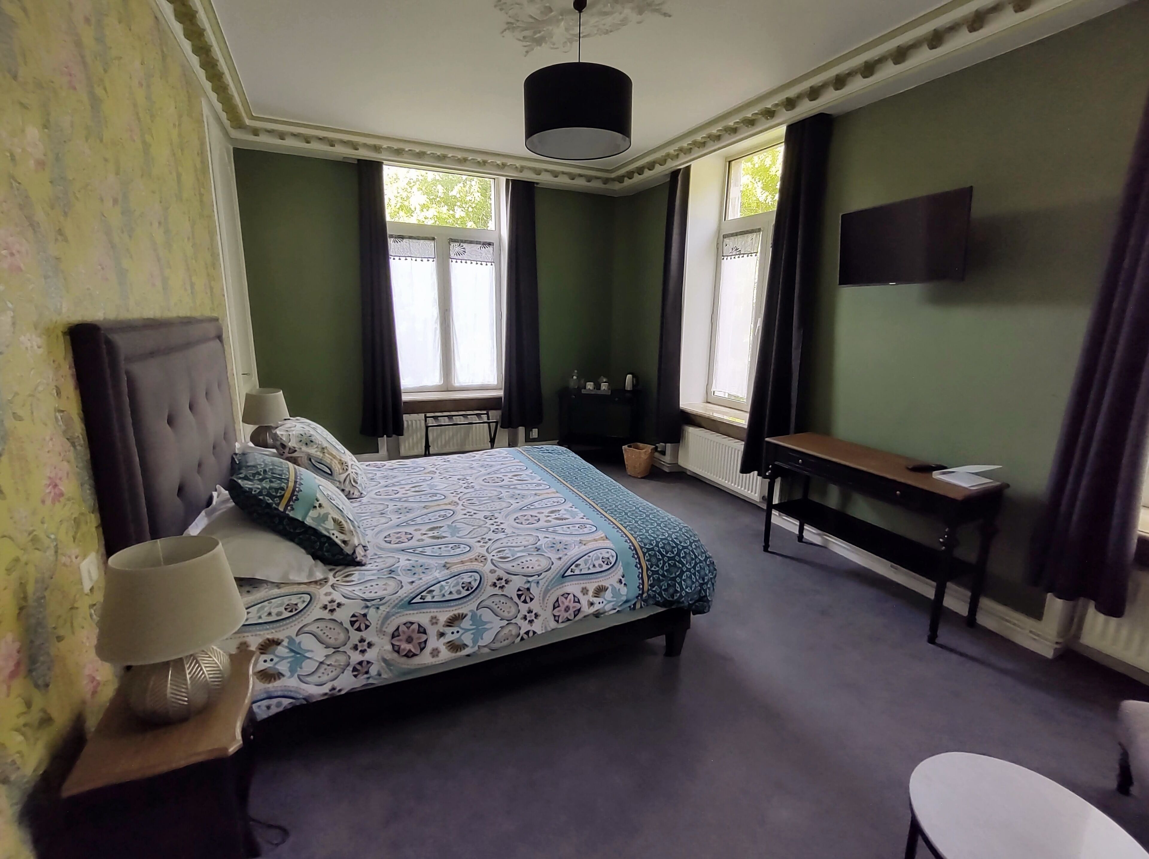 A bedroom with a large bed, a bedside table with a lamp, a desk with a chair, and a TV mounted on the wall.