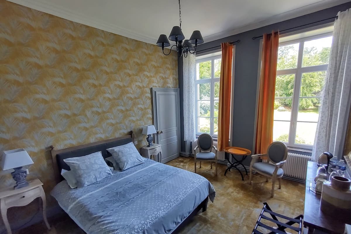 Une chambre à coucher avec un grand lit, une chaise, une petite table et une fenêtre avec des rideaux.