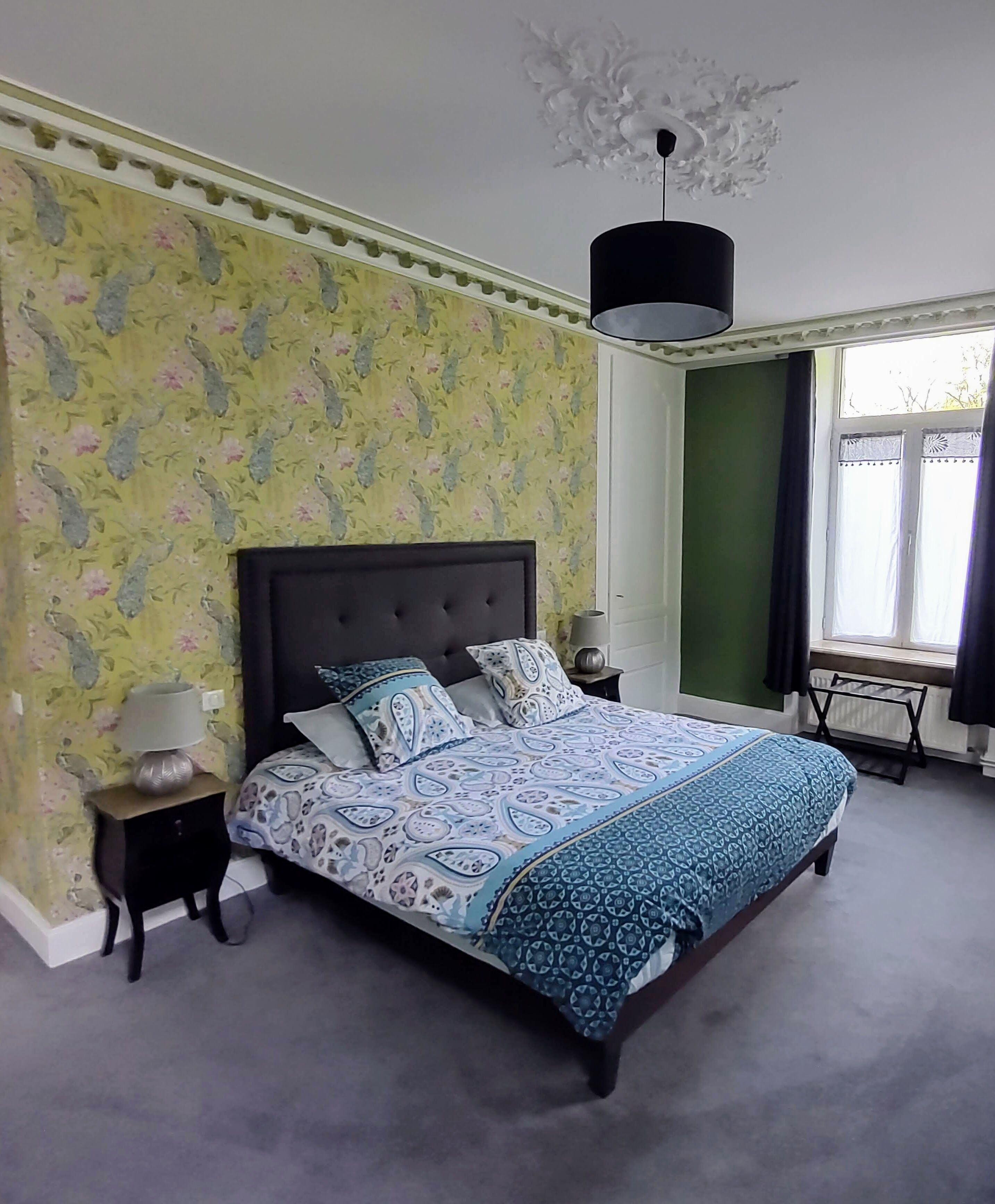 Une chambre avec un papier peint à motifs floraux, une tête de lit capitonnée, une couvre-lit à motifs, une table de chevet avec une lampe et une fenêtre avec des rideaux.