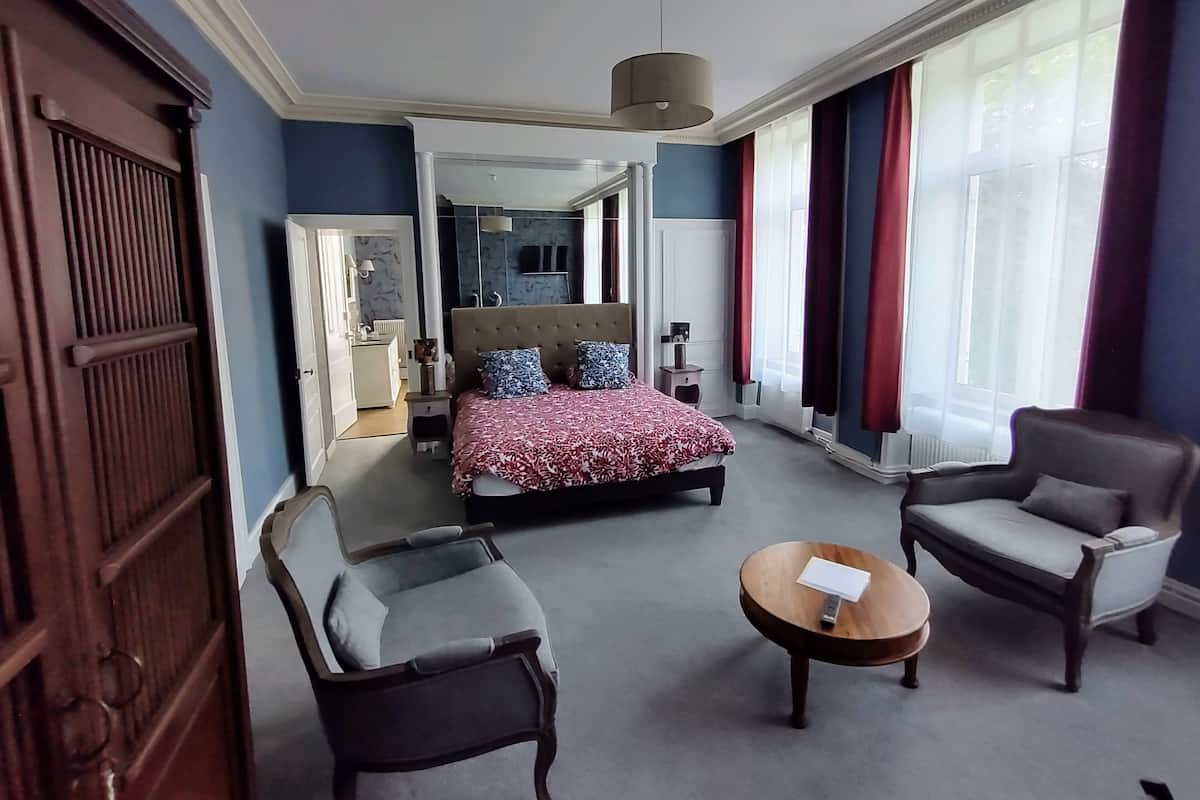 Une chambre d’hôtel avec un grand lit, deux fauteuils, une petite table en bois et une vue sur le paysage urbain, visible à travers de grandes fenêtres.