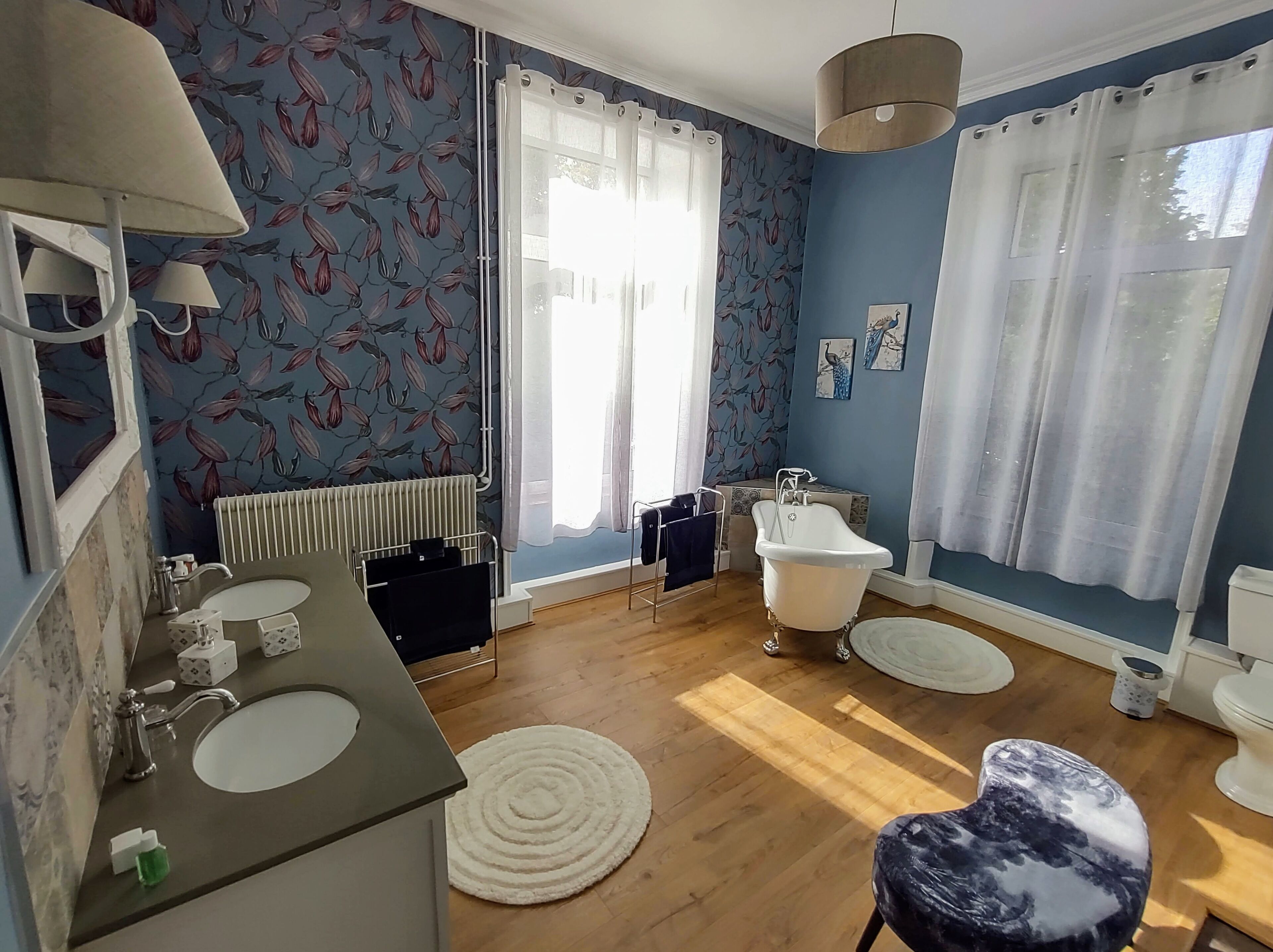 Un baño con bañera, lavamanos e inodoro. Cuenta con dos ventanas con cortinas, un radiador y un espejo.