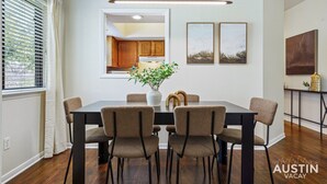 A dining area with a black table, four chairs, a vase with flowers, and a view of the kitchen through a window. - Modern Haven with King Suite + Impeccable Location (Austin)