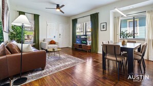 A modern living room with a sofa, coffee table, and dining area. There is a ceiling fan, a TV, and a window with curtains. - Modern Haven with King Suite + Impeccable Location (Austin)