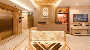 Een hotel receptie met een marmeren receptiebalie, het logo van Bestier Hotel, een receptie counter, een televisie en een wandplank met decoratieve objecten.