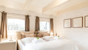 Un letto rifatto con lenzuola e cuscini bianchi, un comodino con una lampada e una finestra con tende.
