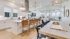 A spacious kitchen with white cabinetry, a large island with bar stools, and a dining area with a long table. - Classic Coastal Charm w/ Modern Style & Amenities @Cinnamon Shore South! (Port Aransas)