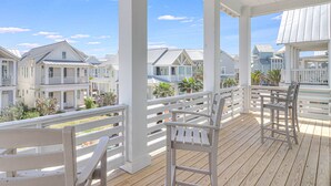 Une terrasse en bois, dotée de rampes et de chaises blanches, qui offre une vue sur une rangée de maisons de plage aux façades blanches et aux toits bleus.