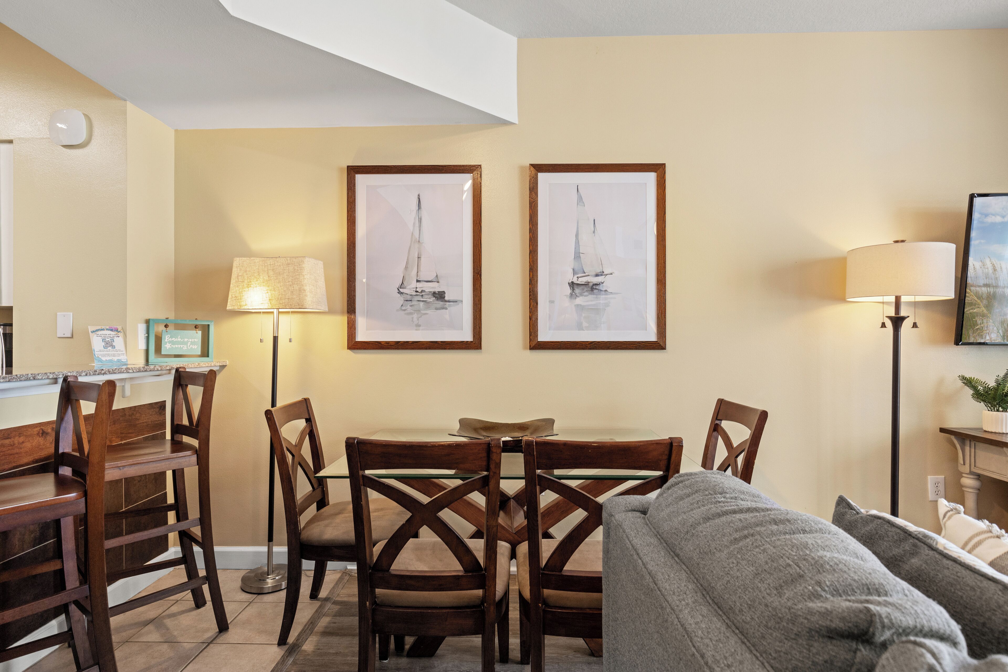 Un comedor con una mesa de vidrio, sillas de madera y dos cuadros enmarcados de veleros en la pared.