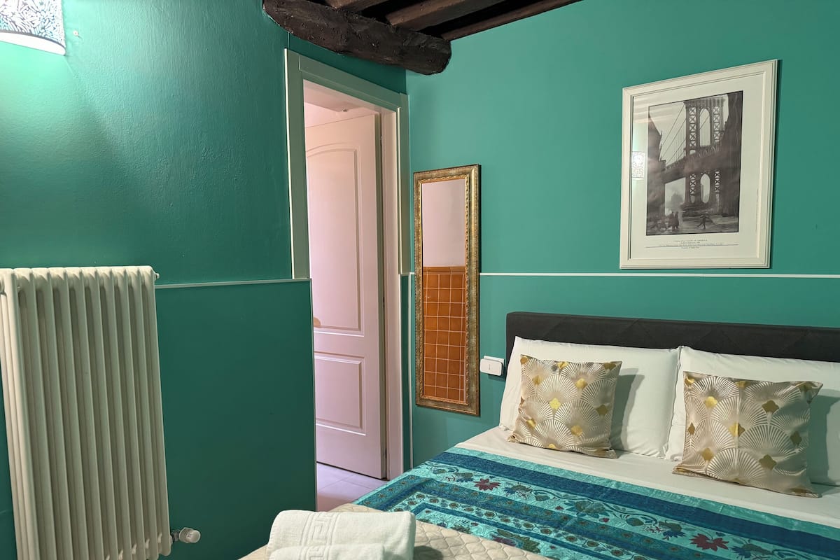 A room with teal walls, a bed with patterned pillows, a framed picture, and a radiator.