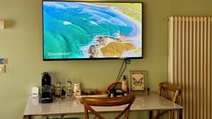 Quarto com televisão de tela plana exibindo uma cena de praia, uma mesa com diversos objetos e uma cadeira.