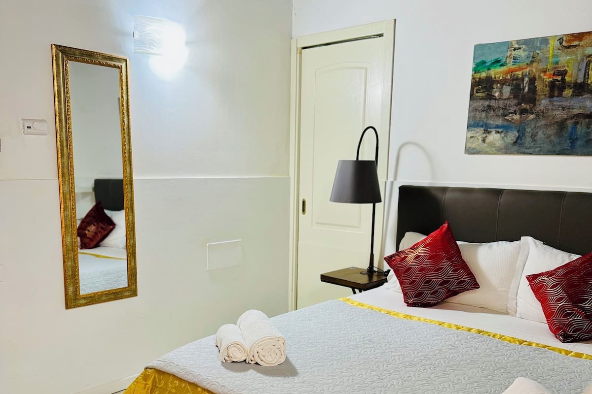 A neatly made bed with a white and gold bedspread, two rolled towels, and red patterned pillows. A black lamp is on a bedside table, and a painting hangs on the wall.