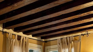 Quarto com duas janelas, cada uma com cortinas bege amarradas, vigas de madeira no teto e luminária fixada na parede.