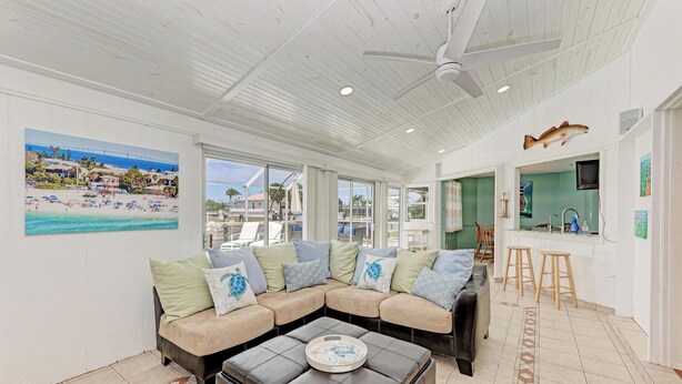 A spacious living area with a large sectional sofa, a coffee table, and a ceiling fan. There is a kitchen area with a bar and stools, and a large window providing a view of the beach. - Hometown Paradise- Canalfront home with dock and heated pool  (Bradenton)
