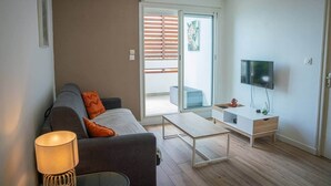 A modern living room with a sofa, a coffee table, a TV, and a sliding glass door leading to an outdoor area. - Bel Appartement vue mer - Mafat'appart (La Possession)