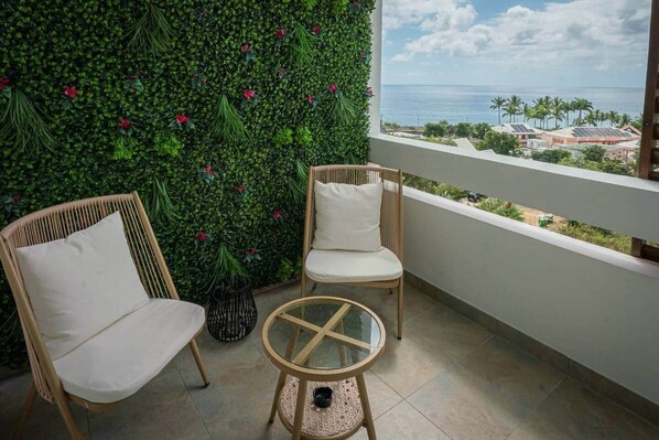 Un balcón con dos sillas de mimbre y una mesa de vidrio, una pared verde con flores rojas, y vista al océano y edificios.