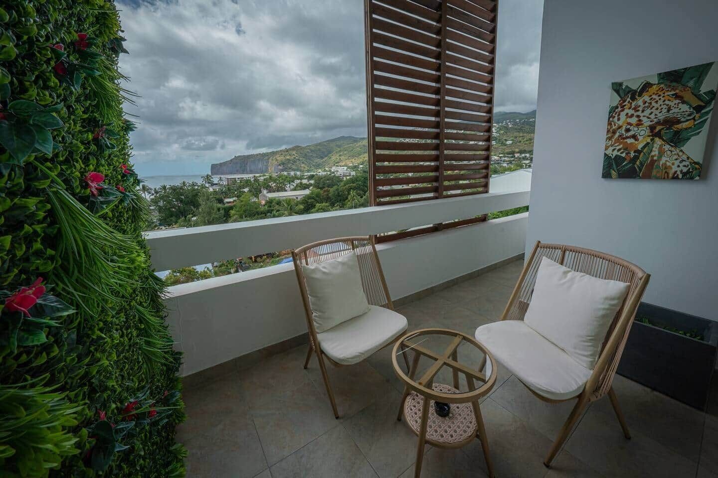 A balcony with two wicker chairs, a view of a coastal landscape, and a green wall with plants.