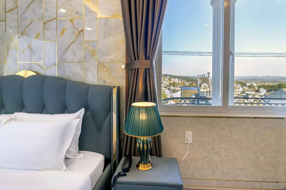 A bedroom with a bed, a nightstand, a lamp, a window with a view, and a marble-patterned wall.