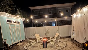 Ein Garten mit Feuerstelle und Liegestühlen, umgeben von einem weißen Zaun und einem hellblauen Gebäude.