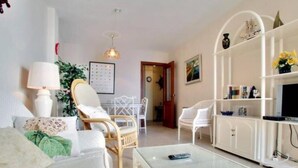 Una sala de estar con un sofá, una silla de mimbre, una mesa de centro de vidrio y un aparador blanco con estantes.