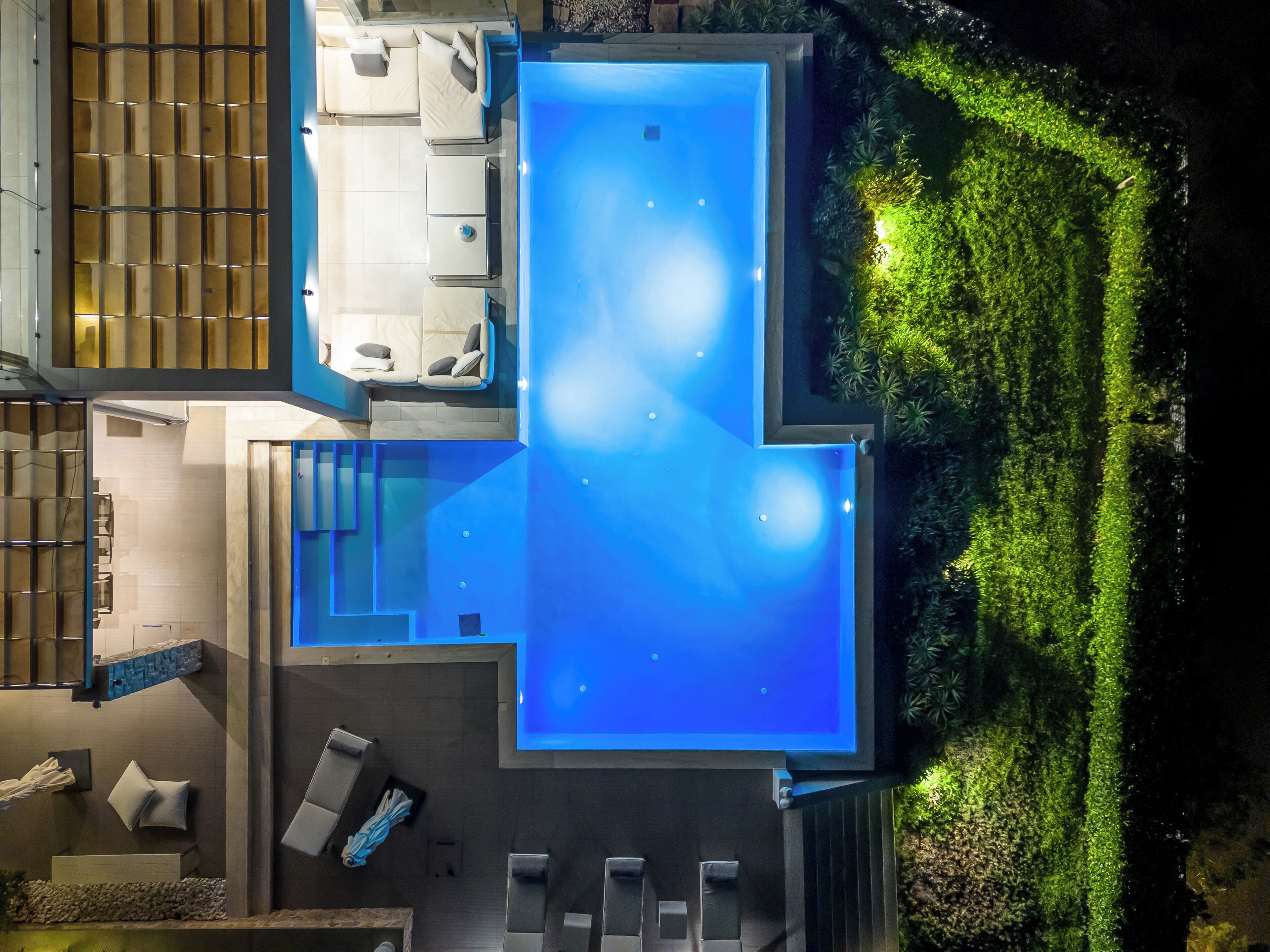 A modern outdoor swimming pool with a geometric design, surrounded by a landscaped garden and a seating area with lounge chairs.