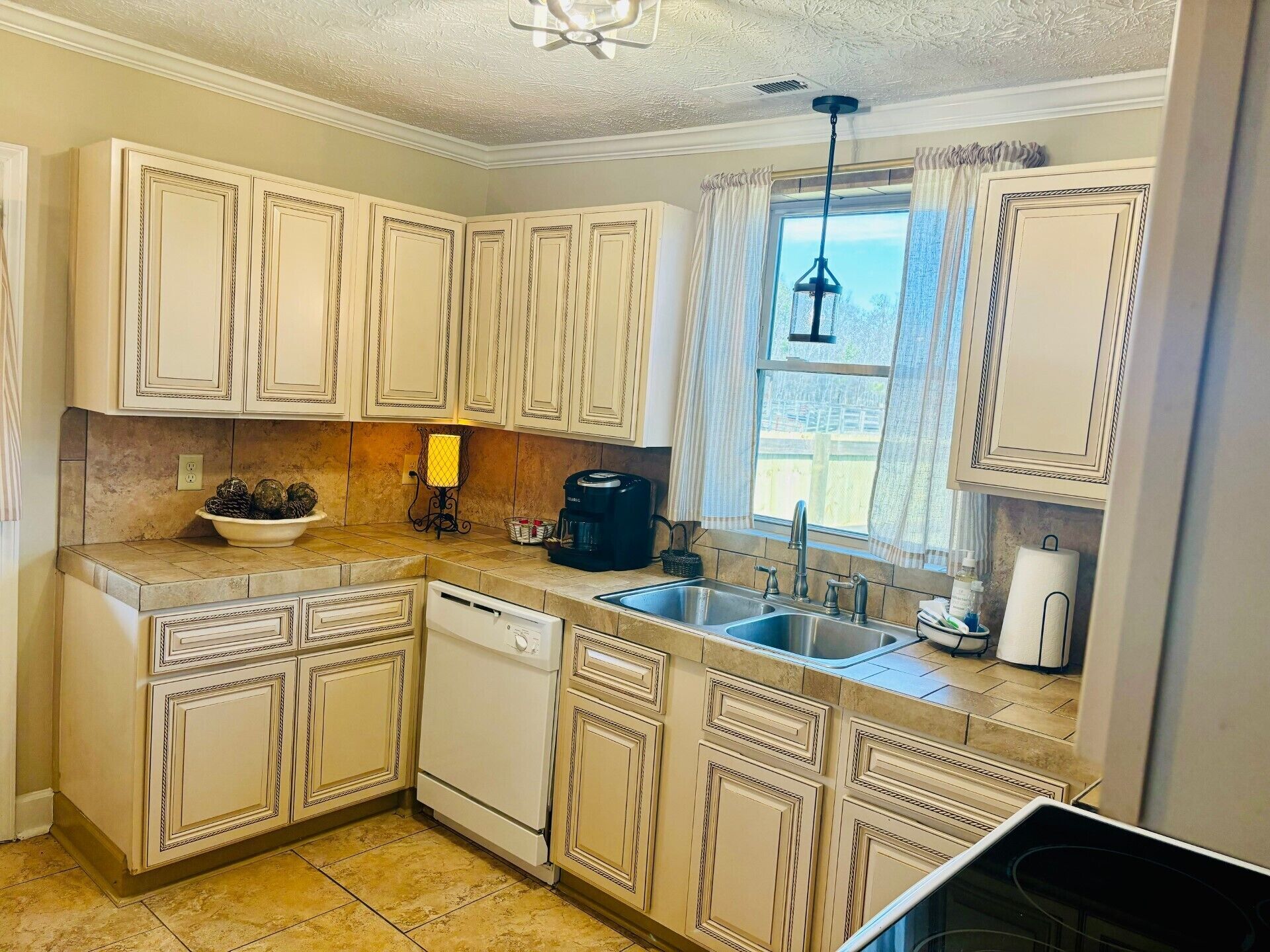 A kitchen with white cabinetry, a dishwasher, a sink, a coffee maker, and a window with curtains.