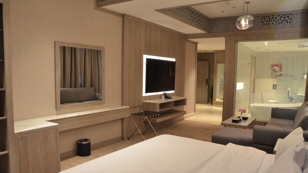A modern hotel room with a large bed, a TV mounted on a wall, a seating area, and a bathroom visible through a glass door.
