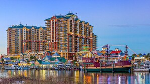 A waterfront hotel with multiple balconies, a marina with boats, and a variety of colorful buildings along the shore. - Beach Vibes, Beachfront Penthouse, Pelican Beach Resort (Destin)