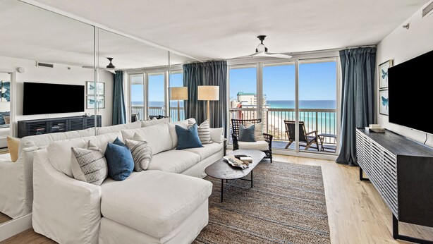 A modern living room with a large sofa, a coffee table, and a TV. There is a balcony with a view of the sea. - Beach Vibes, Beachfront Penthouse, Pelican Beach Resort (Destin)