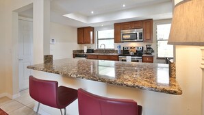 Una cocina moderna con encimeras de granito, armarios de madera y microondas integrado. Hay dos taburetes rojos y un ventanal que proporciona luz natural.