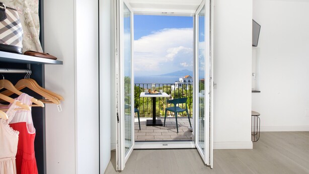 Une porte ouverte donnant sur un balcon équipé d’une table et de chaises, avec vue sur la mer, et un plancher en bois à l’intérieur.