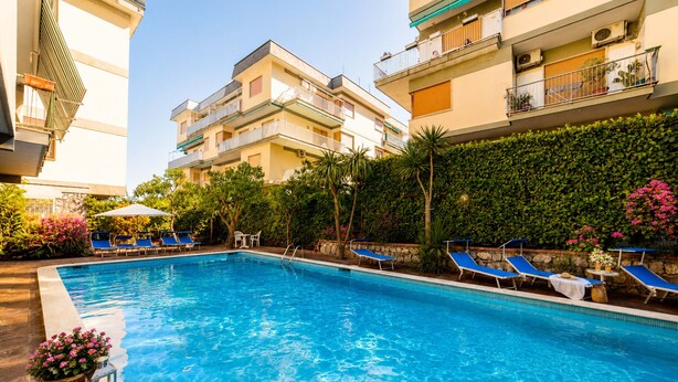 A swimming pool with lounge chairs, a stone wall, and a multi-story building with balconies. - AMORE RENTALS - Appartamento Sally with Terrace and Partial Sea View (Sorrento)