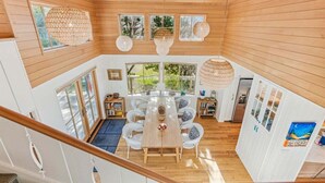 A spacious dining area with a wooden table and chairs, surrounded by large windows offering views of greenery.