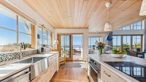 A modern kitchen with wooden flooring, stainless steel appliances, and large windows offering ocean views.