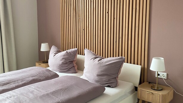 Ein modernes Schlafzimmer mit Bett, Kissen, einem Nachttisch, einer Lampe und einer Wandpaneele aus Holz.