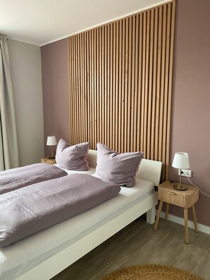 A modern bedroom with a bed, pillows, a nightstand, a lamp, and a wooden panel wall. - Ferienwohnung Nahjkamer im Ortskern von Borkum (Borkum)