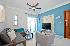 A living room with a sofa, armchair, TV, ceiling fan, and a door with a sign.