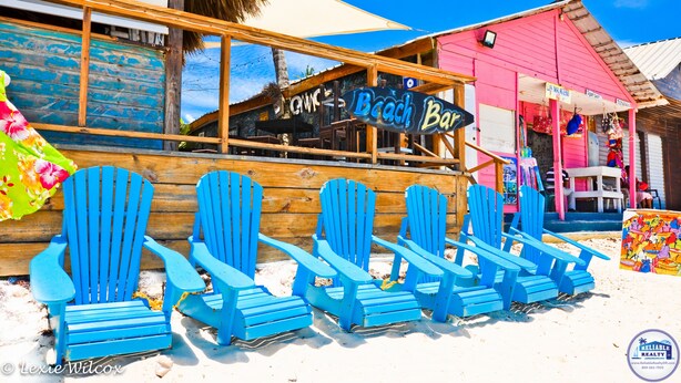A beach bar with a sign reading "Beach Bar" and several blue Adirondack chairs lined up outside.