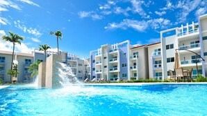 A swimming pool with a water fountain, surrounded by modern buildings with balconies and palm trees. - Beautiful 2-bedroom 3rd level condo with private rooftop terrace  (Punta Cana)