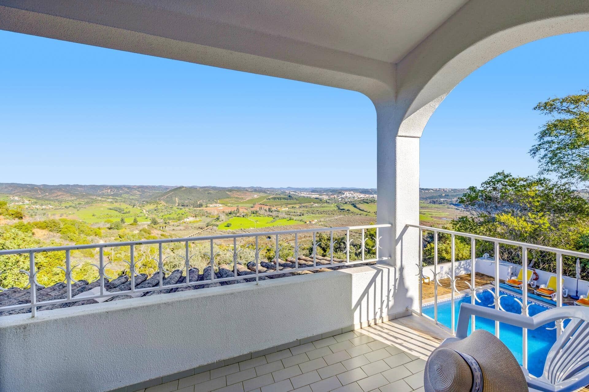 A balcony with a pool, white railing, and a view of a green landscape.