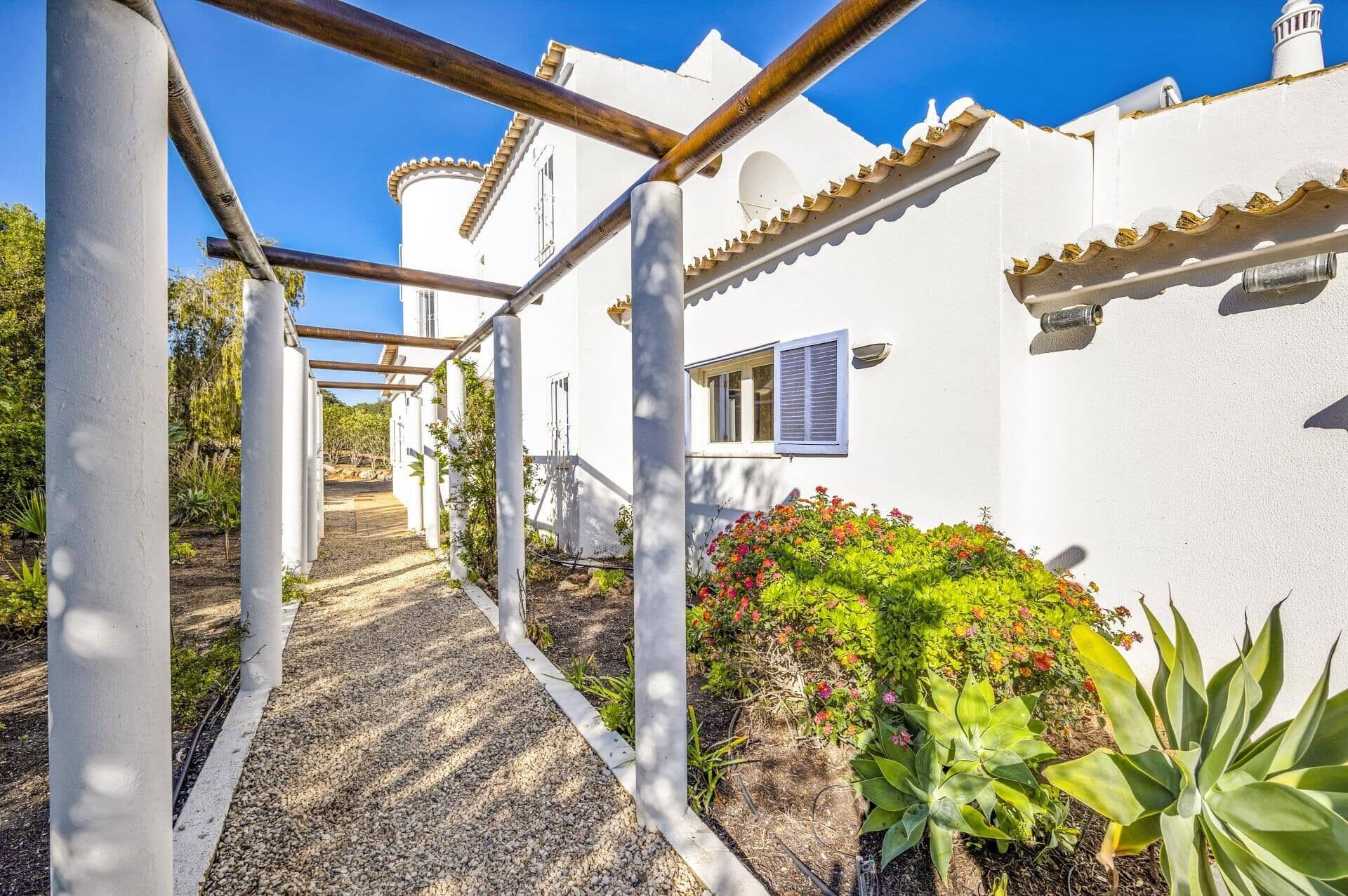 A pathway leading to a white building with a window and a door, surrounded by plants and a clear blue sky.