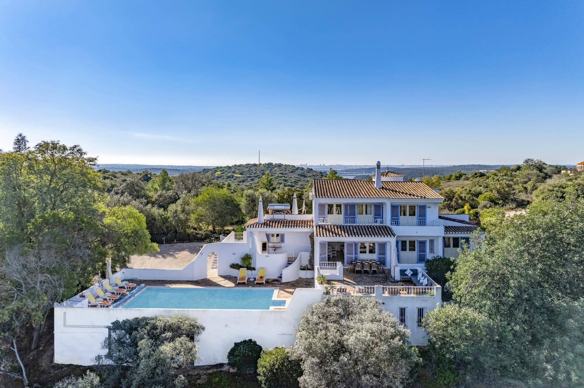 A large, multi-story house with a swimming pool, surrounded by lush greenery and hills.