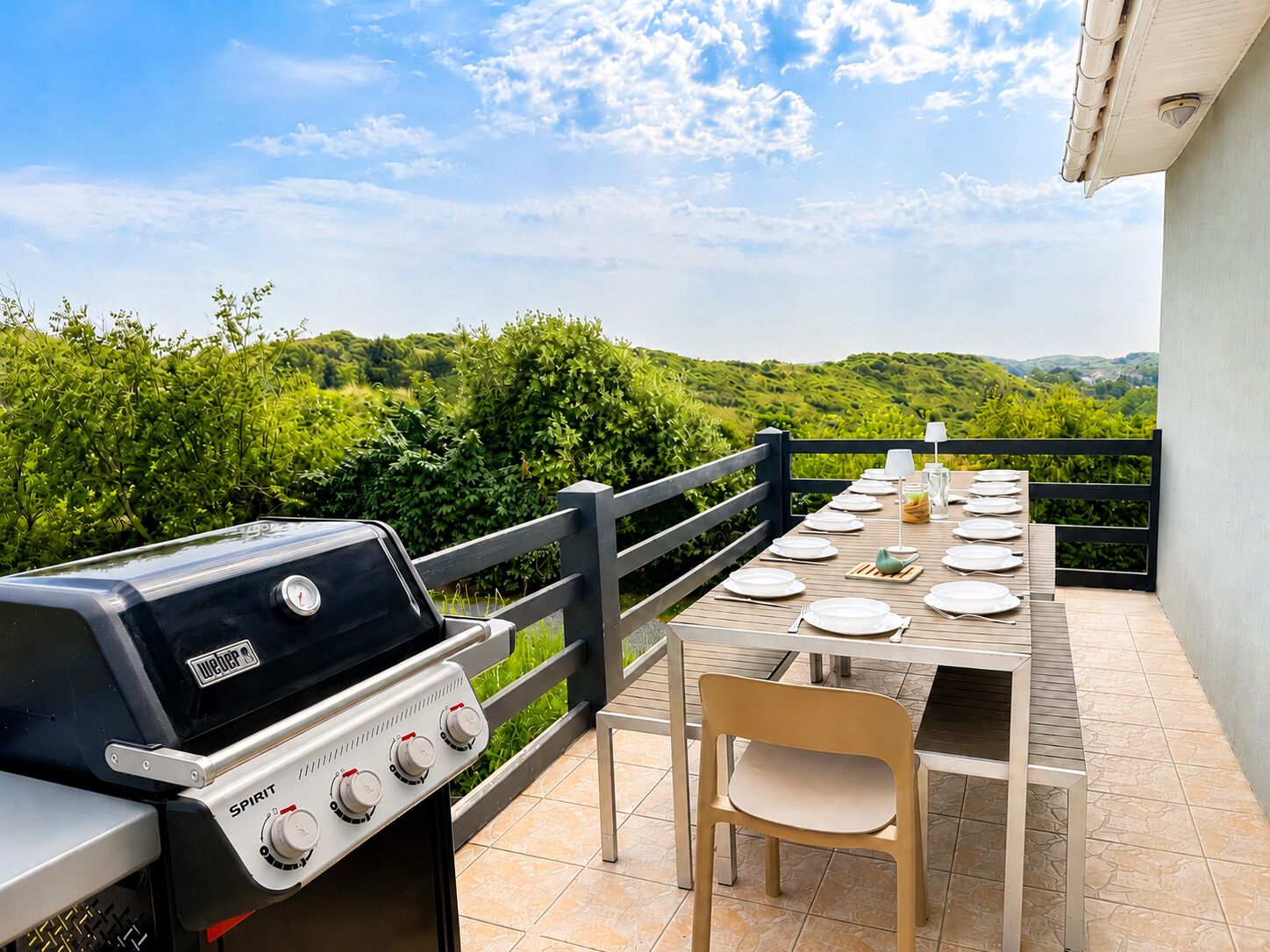 An outdoor dining area with a grill, a table set for four, and a view of greenery.