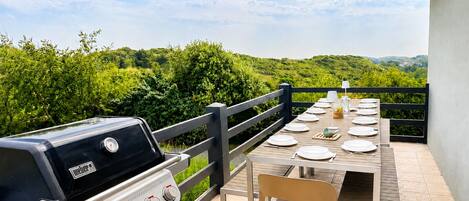 An outdoor dining area with a grill, a table set for four, and a view of greenery.
