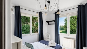 Ein Schlafzimmer mit Bett, Sessel, Schreibtisch und großen Fenstern mit Vorhängen.