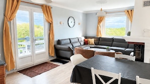 Ein modernes Wohnzimmer mit einer Couch, einem Esstisch, Stühlen, großen Fenstern mit Vorhängen, einer Uhr und einem Kamin.